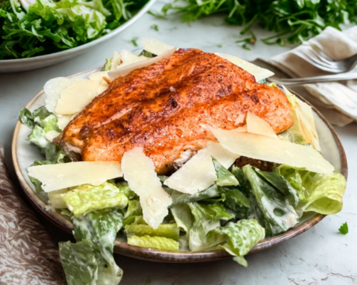 A plate of seasoned salmon atop a fresh Caesar salad, garnished with shaved Parmesan and surrounded by leafy greens.