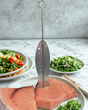 A metal fish Fishtastic gauge inserted into a slab of pink fish, with fresh salads in the background on a marble countertop.